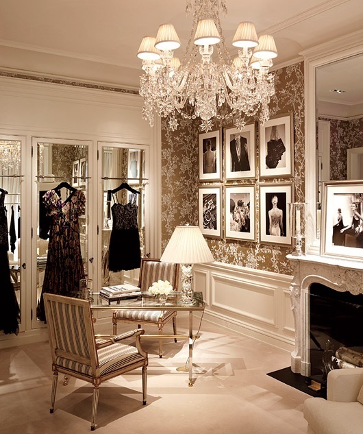 a beautiful and refined closet with paneling and gold printed wallpaper, mirror closets and a fireplace, a glass table and a couple of striped chairs