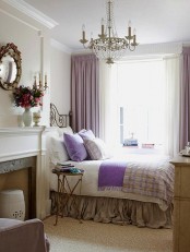 a French country bedroom with a non-working fireplace, a forged bed, some plaid and lavender bedding, a crystal chandelier and a mirror