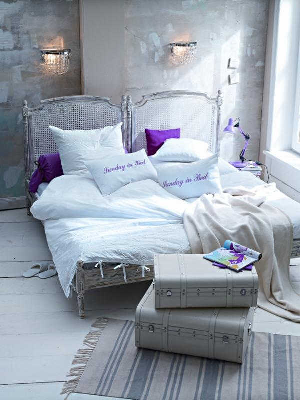 a whitewashed bedroom with brushstrokes on the wall, a whitewashed rattan bed, suitcases and some white and purple bedding