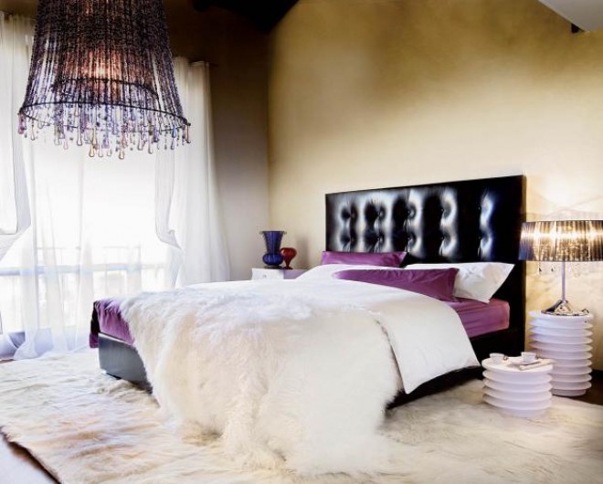 a tan bedroom with a black upholstered bed, purple and white bedding, table lamps and a purple crystal chandelier for colorful accents