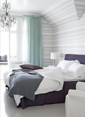a striped bedroom with aqua curtains, a crystal chandelier and a purple upholstered bed plus purple pillows for a color statement