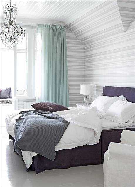 a striped bedroom with aqua curtains, a crystal chandelier and a purple upholstered bed plus purple pillows for a color statement