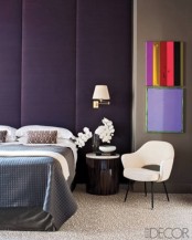 a purple and tan bedroom with a black bed, a dark stained nightstand, colorful artworks and a neutral chair is very refined and chic