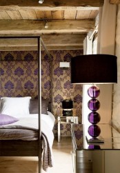 a refined bedroom with tan and purple printed wallpaper, a rough wooden ceiling, a table lamp with a purple ball base and a mirror dresser