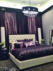 an exquisite bedroom in neutrals and purple, with purple curtains, a white upholstered bed and purple bedding, a pendant lamp with crystals