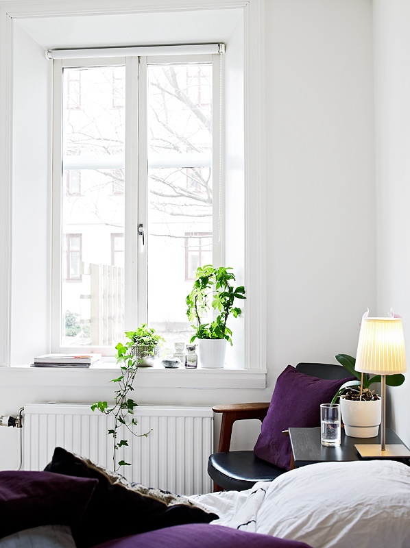 a neutral Scandinavian bedroom with a black leather chair, purple bedding and some potted plants and a table lamp