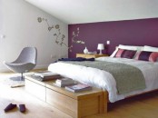 an attic bedroom with light-stained furniture and a grey chair, with purple and white bedding and some floral decor