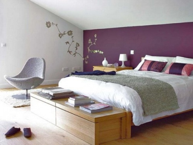 an attic bedroom with light stained furniture and a grey chair, with purple and white bedding and some floral decor