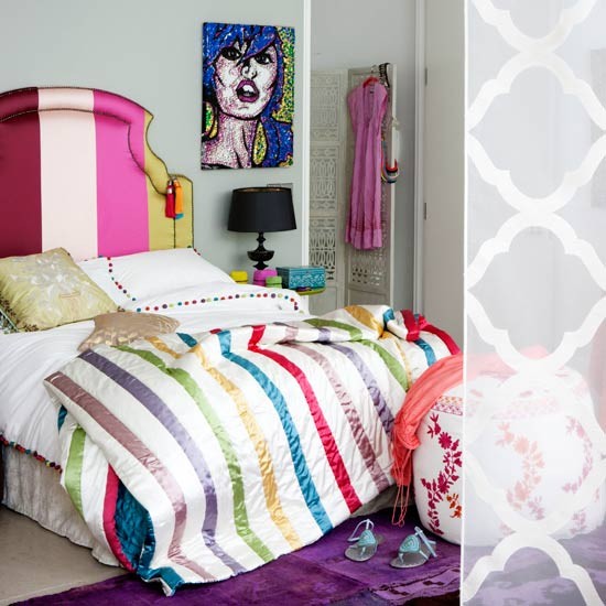 a grey bedroom made bright with a bed with a rainbow striped headboard and matching bedding, a purple rug, a floral print pouf and a bold artwork