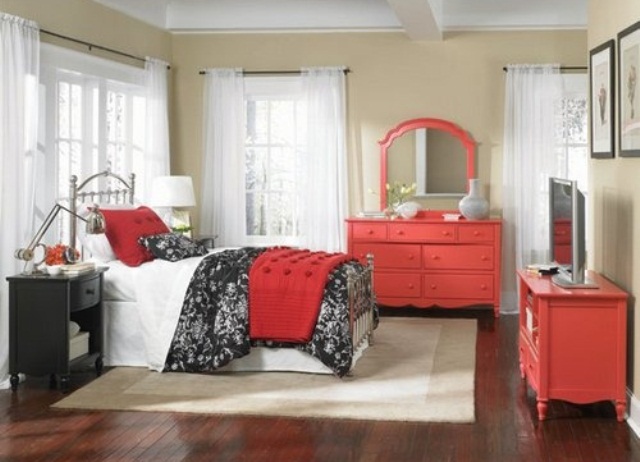 a beige bedroom with a metal bed, a black dresser, red dressers, bold and printed bedding, table lamps and blooms