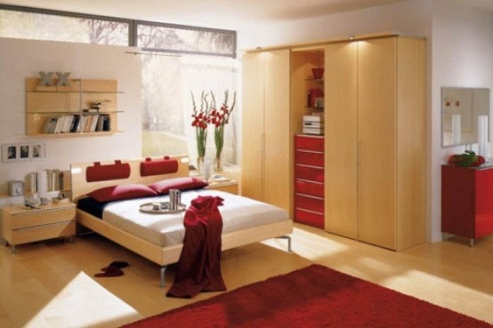 a light-colored bedroom with a glazed wall, a light-stained bed and a wardrobe, a red chest of drawers, red bedding and a red rug