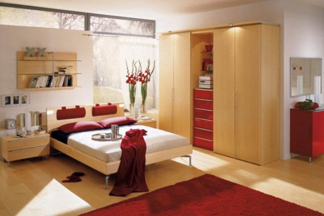 a light colored bedroom with a glazed wall, a light stained bed and a wardrobe, a red chest of drawers, red bedding and a red rug
