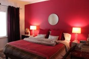 a bedroom with a red accent wall, a bed with red and neutral bedding, burgundy curtains and mirror nightstands