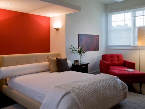 a modern neutral bedroom with a red accent wall, a bed with neutral bedding, a red chair with a footrest, lamps and sconces