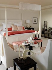 a refined neutral bedroom with a frame canopy bed, an upholstered bench and neutral chairs, a black side table and a printed chair, red bedding as an accent
