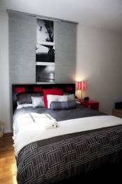 a small bedroom with a grey accent wall, some black and white artwork, a red nightstand and red pillows and grey and white bedding