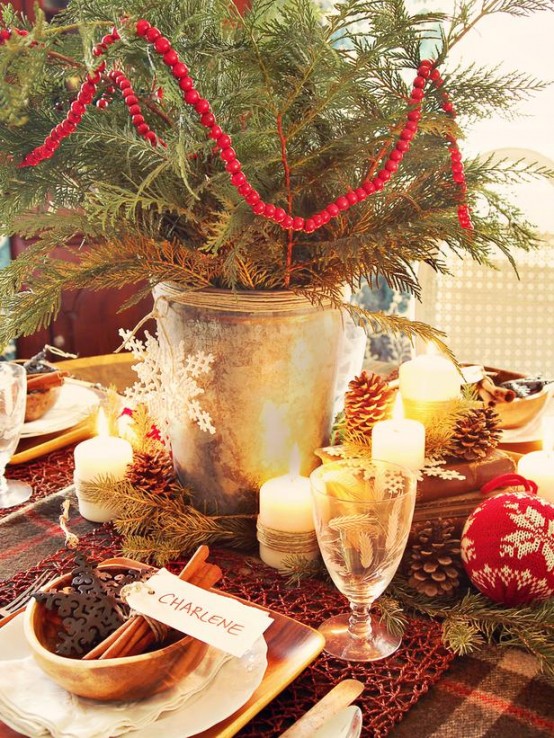 a cozy rustic Christmas tablescape with a plaid tablecloth, red yarn placemats, pinecones, candles and a Christmas tree in a bucket, with a red cranberry garland