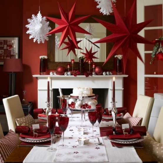 a stylish red and white Christmas dining room with a non-working fireplace, a mirror over it, a table with neutral chairs, red and white paper stars hanging over the tabl and some red tableware