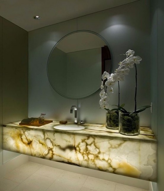 a luxurious contemporary bathroom with grey walls, a lit up onyx slab vanity and a large round mirror