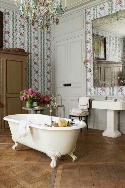 a lovely vintage bathroom with floral wallpaper, a wardrobe a white clawfoot bathtub, a free-standing sink, an oversized mirror and a gorgeous parquet floor