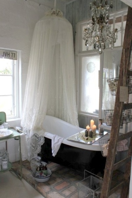 a neutral vintage bathroom with a black clawfoot bathtub and a canopy over it, a crystal chandelier and a ladder, a free-standing sink and some vintage signs and accessories