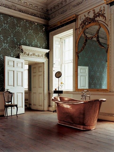 a vintage bathroom with green printed wallpaper, fantastic molding, an oversized mirror in an ornated frame, a copper bathtub, a vintage chair and a side table