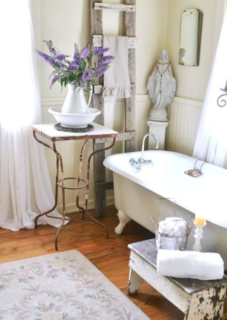 a neutral vintage countryside bathroom with neutral paneling, a tan clawfoot bathtub, a wooden ladder for towels and a side table plus a bench for storage