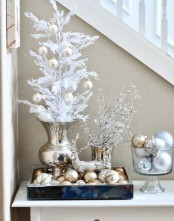 a gold urn with a white Christmas tree with gold ornaments, a tray with gold and gold glitter ornaments, a bowl with them and icy branches