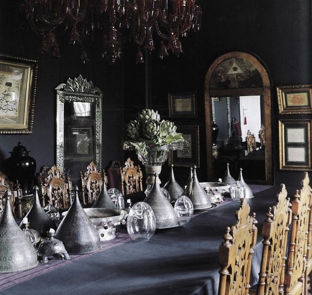 a beautiful and moody Gothic dining room with black walls, a gorgeous red chandelier, mirrors and artworks, a long table with cones and carved wooden chairs