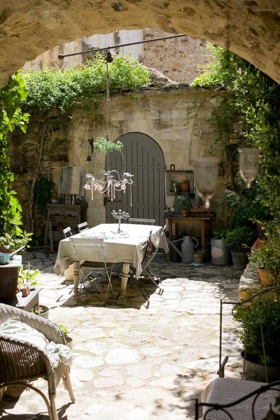 a Provence inner courtyard with a metal table and vintage folding chairs, wicker furniture, greenery and vintage lamps and a chandelier