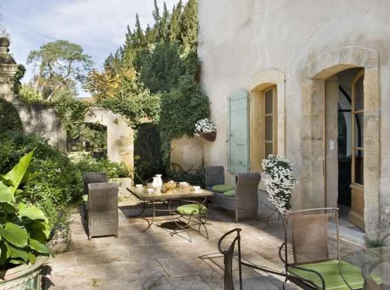 a beautiful Provence terrace with metal and glass tables, wicker chairs with green cushions, lots of greenery and blooms around