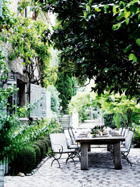 a refined Provence terrace with a dining space, a stained table, black and white chairs with comfortable upholstery and some greenery and trees around