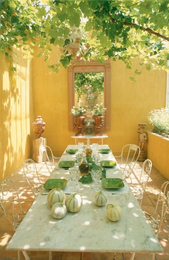 a Provencal terrace done in sunny yellow, with a long table of metal and stone and metal chairs, greenery around and some vintage urns