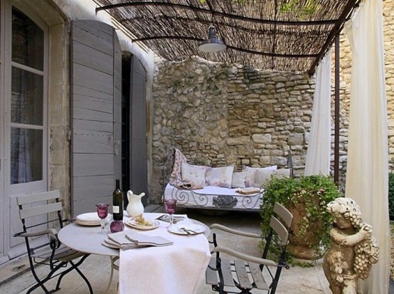 a chic Provence terrace a round metal table and metal and wood chairs, a forged daybed with neutral cushions and pillows and potted greenery