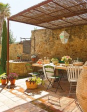 a lovely Provence terrace with a woven roof, a wooden table, wood and metal green chairs, potted blooms and a pendant lantern