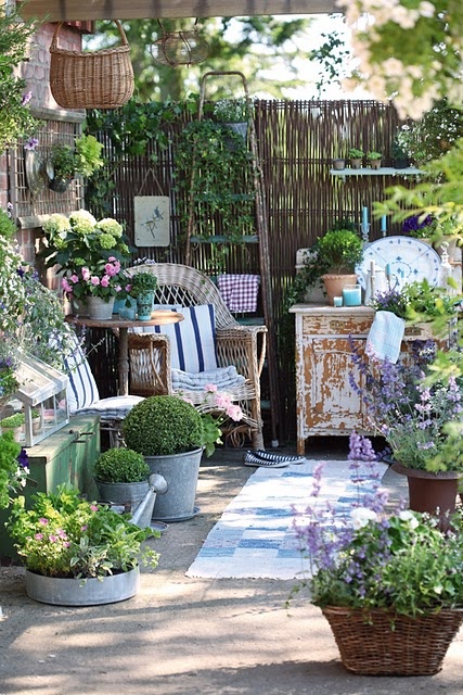 a small Provence-inspired terrace with a wicker chair and a shabby chic cabinet, lots of potted greenery and blooms and some watering cans