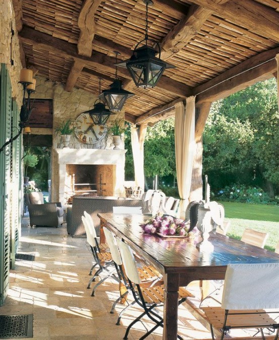 a Provence terrace with a fireplace and some chairs around it, a long stained dining table and neutral chairs, lanterns on the ceiling