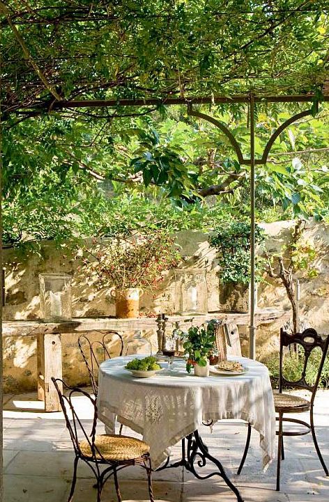 a Provence terrace with lots of greenery that composes a ceiling, a metal table and some mismatching wood and woven chairs and a wooden bench