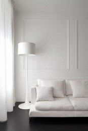 a clean white space with walls with molding, with a white sofa and a floor lamp plus a dark floor that conrasts all the rest
