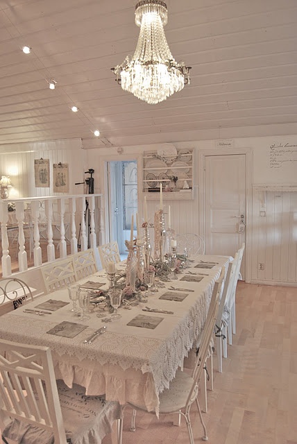 a shabby chic dining space with whitewashed wooden walls and a ceiling, a gorgeous crystal chandelier that highlights the ceiling and refined white furniture plus lots of silver on the table