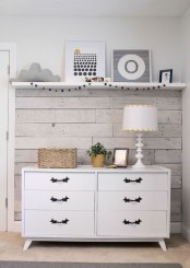 a part of a nursery with whitewashed wood on the wall, a shelf for displaying art, a white dresser and a cool table lamp
