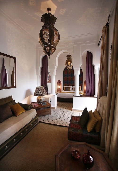 a Moroccan living room with carved wood furniture, pillars, a traditional lantern and a cozy alcove with a bed