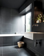 a moody Nordic bathroom fully clad with large grey tiles, with a wall-mounted sink and a tub clad with tiles