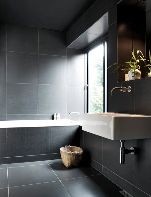 a moody Nordic bathroom fully clad with large grey tiles, with a wall-mounted sink and a tub clad with tiles