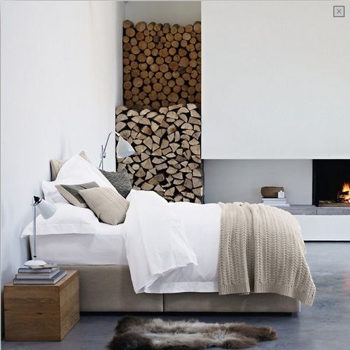 a neutral minimalist bedroom with a bed, nightstands, a fireplace and some firewood stored by it is a stylish space