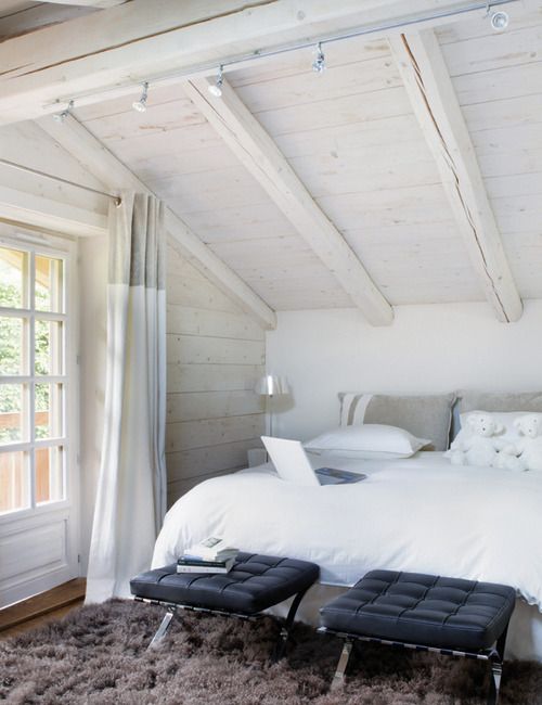 a white attic bedroom with a bed, black leather stools and a brown rug for a drama and a contrast