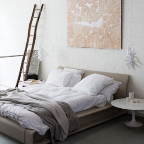 a neutral bedroom with a grey upholstered bed, mismatching nightstands, a ladder, an artwork