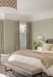 a neutral and muted color bedroom with green walls, a bed with various neutral bedding, a pendant lamp and printed bench