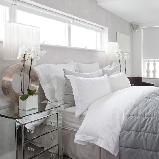 a neutral bedroom with a creamy bed, white and grey bedding, mirror nightstands and potted orchids looks glam and chic