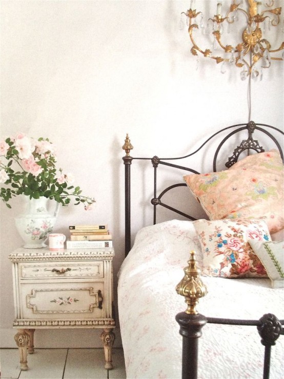 a Provence bedroom in neutrals and pastels, with a metal bed and pastel textiles, a vintage nightstand, blooms, a gold and crystal chandelier